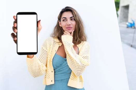 Portrait of beautiful woman in green summer dress on white background natural daylight, pointing on empty white big screen of smartphone, space for advertising