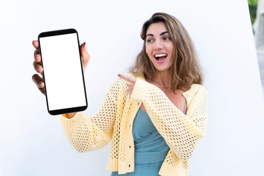 Portrait of beautiful woman in green summer dress on white background natural daylight, pointing on empty white big screen of smartphone, space for advertising