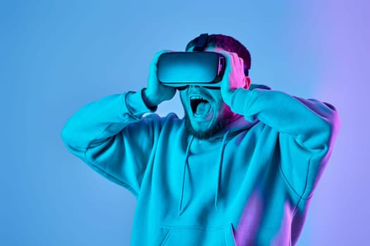 Astonished man in sweatshirt using virtual reality glasses and playing video games on blue background. Neon lighting