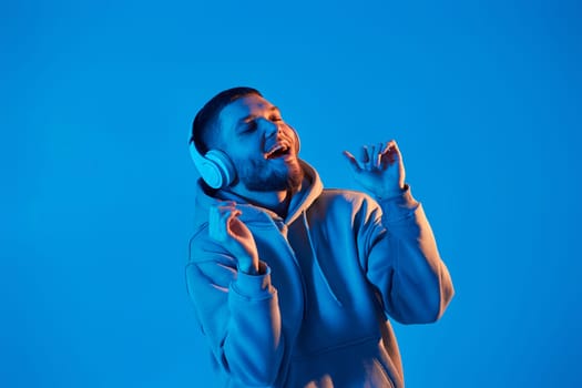 handsome bearded man with headphones in sweatshirt enjoying favorite music on blue neon background. Neon lighting