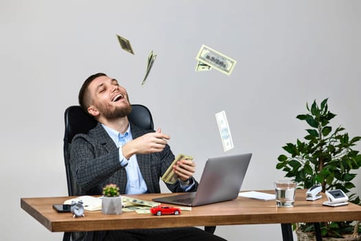 young bearded businessman working on laptop and throwing money while sitting on chair at desk. banknotes fly in the air