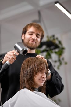 man hairdresser using hair dryer for female hair after washing in the beauty salon.