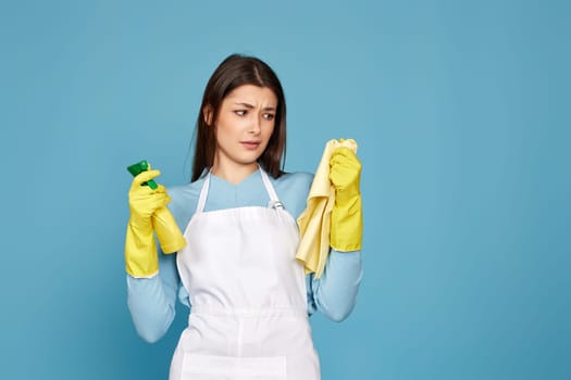 skeptic and nervous, frowning woman in gloves and cleaner apron with cleaning rag and detergent sprayer on blue background.