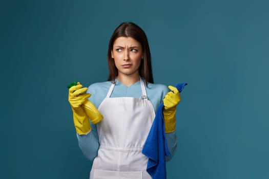 skeptic and nervous, frowning woman in gloves and cleaner apron with cleaning rag and detergent sprayer on blue background.
