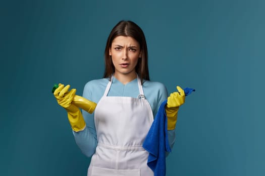 skeptic and nervous woman in gloves and cleaner apron with cleaning rag and detergent sprayer on blue background.