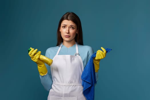 skeptic and nervous, frowning woman in gloves and cleaner apron with cleaning rag and detergent sprayer on blue background.