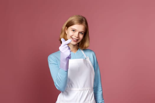 beautiful blonde woman in rubber gloves and cleaner apron showing love gesture on pink background.