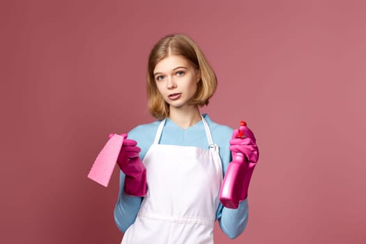 skeptic and nervous, frowning blonde woman in gloves and cleaner apron with cleaning rag and detergent sprayer on pink background.