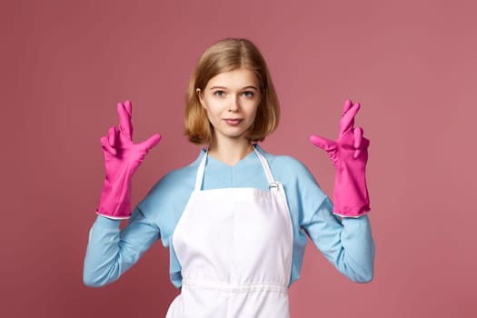 beautiful blonde caucasian girl in rubber gloves with finger crossed gesture on pink background.