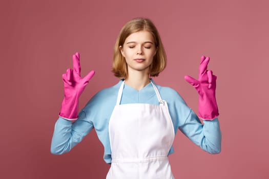 beautiful blonde caucasian girl in rubber gloves with finger crossed gesture on pink background.