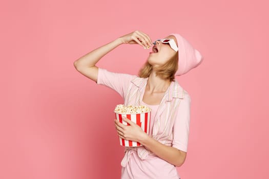 happy blonde woman in 3d glasses eating popcorn on pink background.