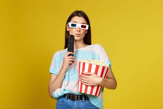 puzzled woman with remote control watching interesting movie and holding bucket of popcorn on yellow background.