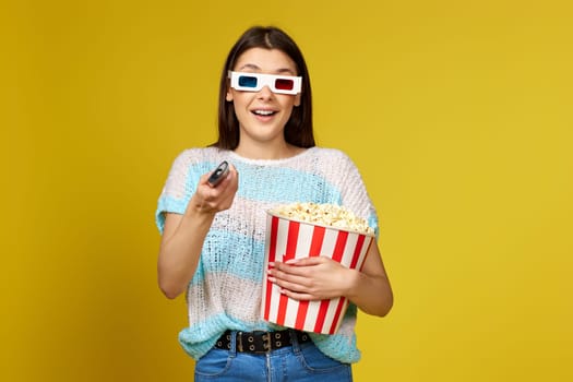 happy woman with remote control watching interesting movie and holding bucket of popcorn on yellow background.