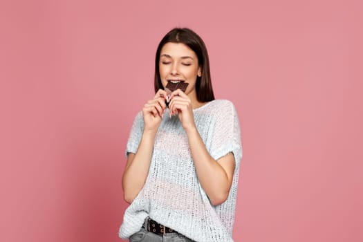 happy woman in sweater biting tasty chocolate bar and looking at camer isolated on pastel pink background