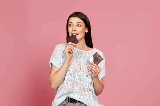 charming happy woman in sweater biting sweet chocolate bar and looking at the side isolated on pastel pink background