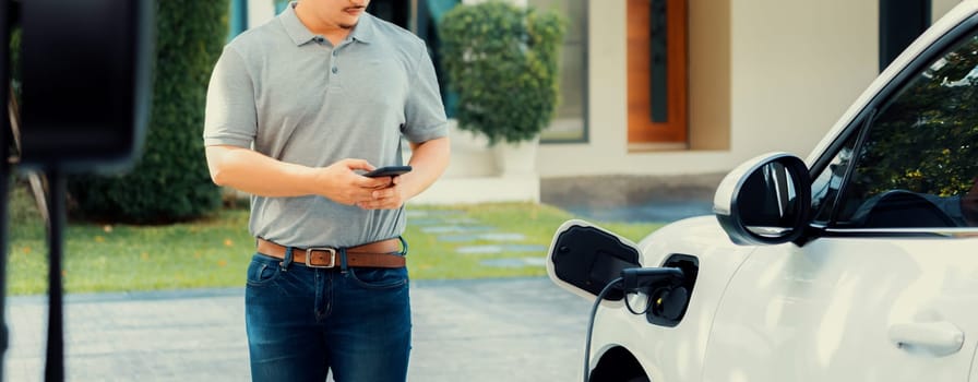 Progressive asian man install cable plug to his electric car with home charging station in the backyard. Concept use of electric vehicles in a progressive lifestyle contributes to clean environment.