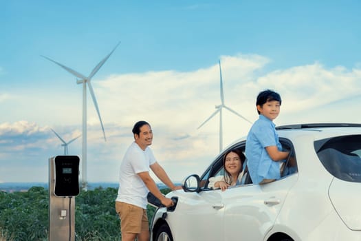 Concept of progressive happy family enjoying their time at wind farm with electric vehicle. Electric vehicle driven by clean renewable energy from wind turbine generator for charging station.