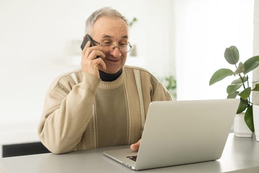 Handsome smiling senior man wearing glasses using mobile phone while sitting at his cozy workplace with laptop at home, retired male chatting with friends in social media, typing on smartphone