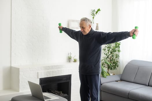 Sport in mature age. Happy middle-aged man doing stretching exercises in front of laptop at home, watching online tutorials, in living room interior.