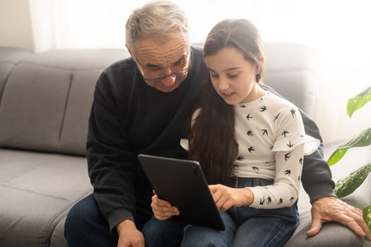 Cute little girl and her handsome grandpa are smiling while sitting on couch at home. Girl is using a tablet