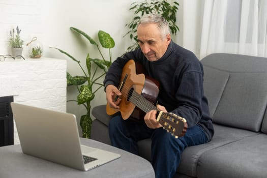 Portrait of senior man in headphones taking online guitar lesson looking at laptop screen. Retired male learning to play guitar watching webinar on computer at home.