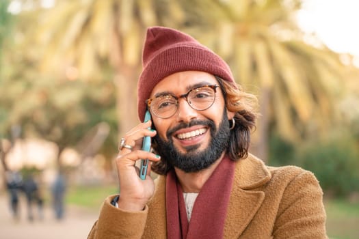 Attractive young caucasian man with glasses and beard talking in the phone outdoors in the park in winter. Happy smiling student call. Technology, education and communication concept.