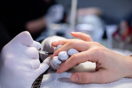 Close-up on the hands of a beauty salon client. In the background the blurred interior of the beauty salon. The beautician wears disposable nitrile gloves. Performing a hybrid manicure on a natural nail plate.