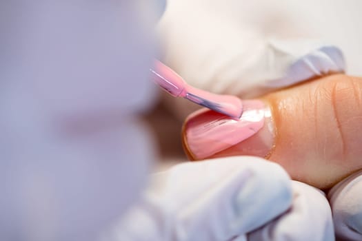 Close-up on the hands of a beautician holding the finger of a client. A woman using the services of a beauty salon. Painting nails with hybrid method. Beautician painting nails with pink polish.