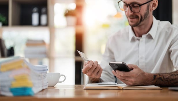 A man reading information from the back of the credit card and typing it on the smartphone to do an online purchase at home. A guy doing an online payment on a cellphone at table in office..