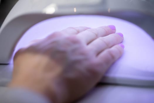 Close-up of a woman's hand. A woman holds her hand inside a lamp that emits ultraviolet light. Performing a manicure using a hybrid method. Using the services of a beauty salon.