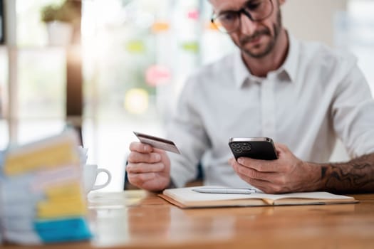 A man reading information from the back of the credit card and typing it on the smartphone to do an online purchase at home. A guy doing an online payment on a cellphone at table in office..