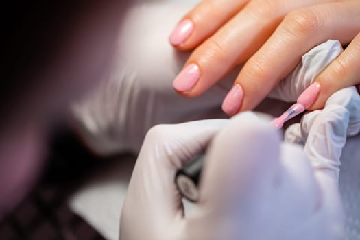Close-up of the hands of a beauty salon client. A woman using the services of a manicurist. The beautician paints the client's nails pink. Applying a layer of hybrid varnish.