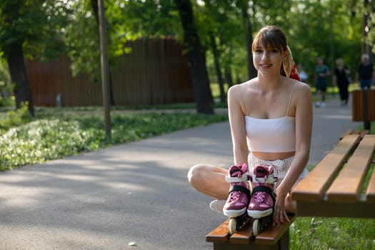 A smiling young woman sits on a bench with her legs crossed. The girl holds rollerblades standing on the bench in front of her. In the background you can see a green park, trees, lawns and an asphalt alley.