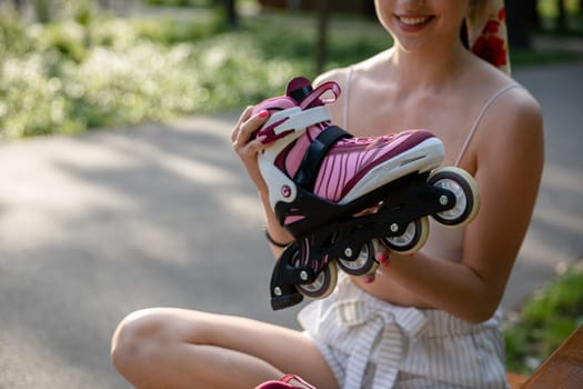Close-up of a rollerblade in shades of gray and purple. A girl holds a rollerblade in her hands. Blurred trees and an asphalt park alley are visible in the background. Sun rays on the asphalt.