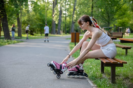A woman improves the fastening of rollerblades while sitting on a bench. A girl puts on rollerblading sports equipment. In the distance, blurry figures of people playing sports and walking down an alley.