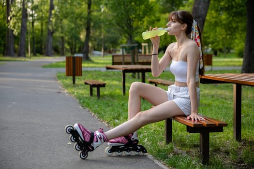 A young woman sits on a bench and drinks from a bottle. Spending time in a green city park. Replenishing electrolytes while playing sports. A hot day in nature.