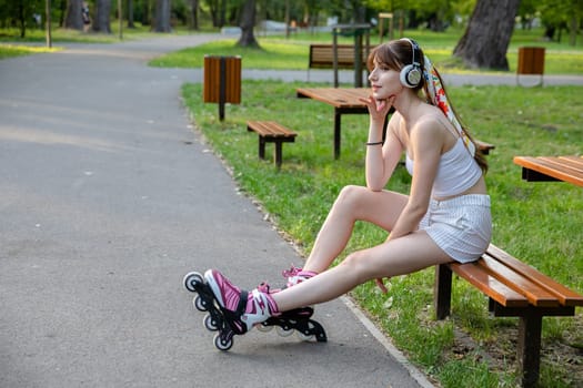An athlete relaxes during training while sitting on a bench. Spending time in a green city park. A young woman dressed in sportswear. The girl is listening to music on wireless headphones.