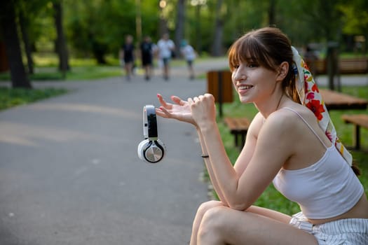 A happy girl sits on a park bench. She is holding wireless headphones in front of her. There are lots of trees and greenery around. In the distance you can see fuzzy figures of people walking.