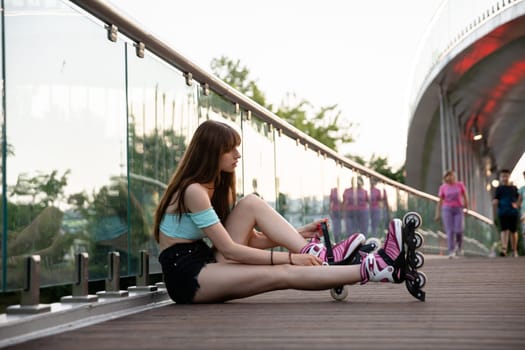 A long-haired girl sits on a bridge and puts on rollerskates. She has a glass railing behind her back. In the background you can see blurred figures of people walking.