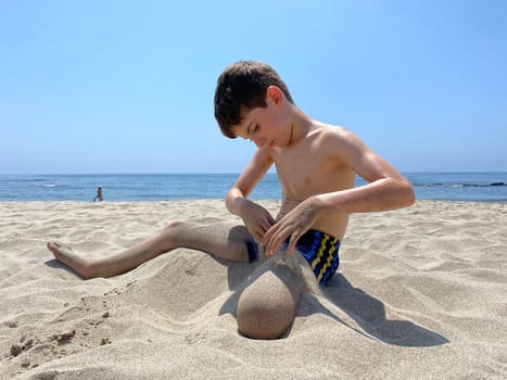 Child sitting on sandy beach pouring sand with his hands over his legs.