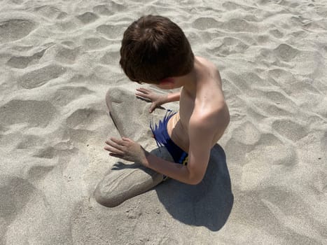 Top view of a Child sitting on sandy beach pouring sand with his hands over his legs