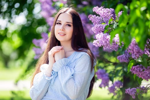 Pretty smiling brunette girl with long hair stands with lilac flowers, in the garden, in sunny day, look at camera. Close up. copy space