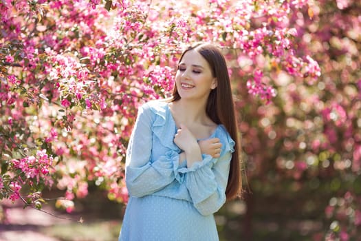 A happy smiling girl, in light blue dress, with dark long hair, standing in a pink blooming garden. Close up
