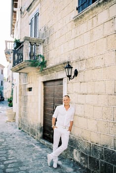 Smiling man in a white shirt stands leaning against an old stone house. High quality photo