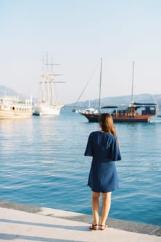 Woman in a blue dress stands on the pier and looks at the ships. High quality photo