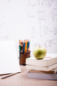 Green apple on pile of books next to a notebook and pencils on table with a blurred white board in the back. School concept