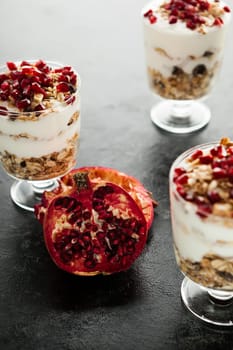 Half of pomegranate lying in the middle on three glasses with muesli on dark wooden background