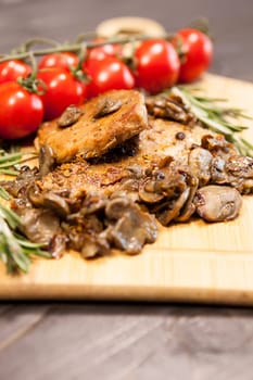 Close up on wooden board with grilled mushrooms and pork steak next to cherry tomatoes on a wooden table