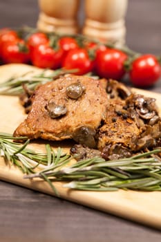 Delicious pork steak on wooden board next to a branch of oregano and cherry tomatoes. Two shakers with salt and pepper are blurred in the background