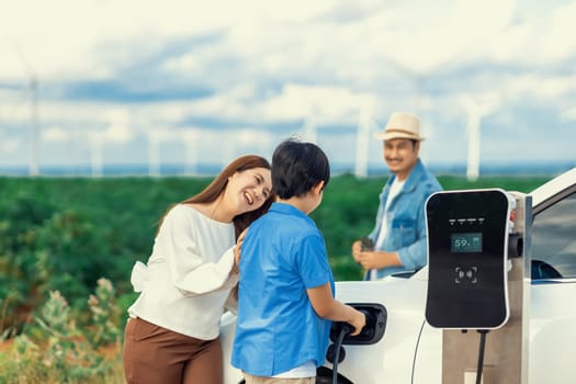 Concept of progressive happy family enjoying their time at wind farm with electric vehicle. Electric vehicle driven by clean renewable energy from wind turbine generator for charging station.